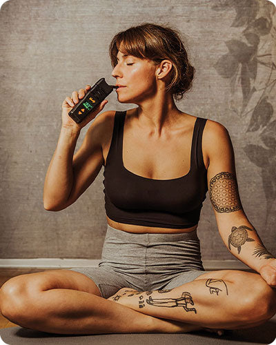 Mujer sentada con el vaporizador VENTY de Storz & Bickel en la mano y la boquilla del VENTY en los labios, frente a una pared texturizada con motivo floral